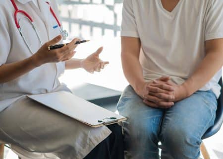A doctor in a white coat consulting a patient in a white T-shirt and blue jeans.