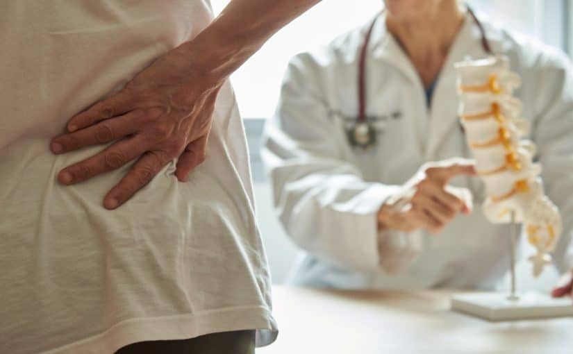 A patient holding his back in pain in front of a doctor gesturing toward a model of a spine.